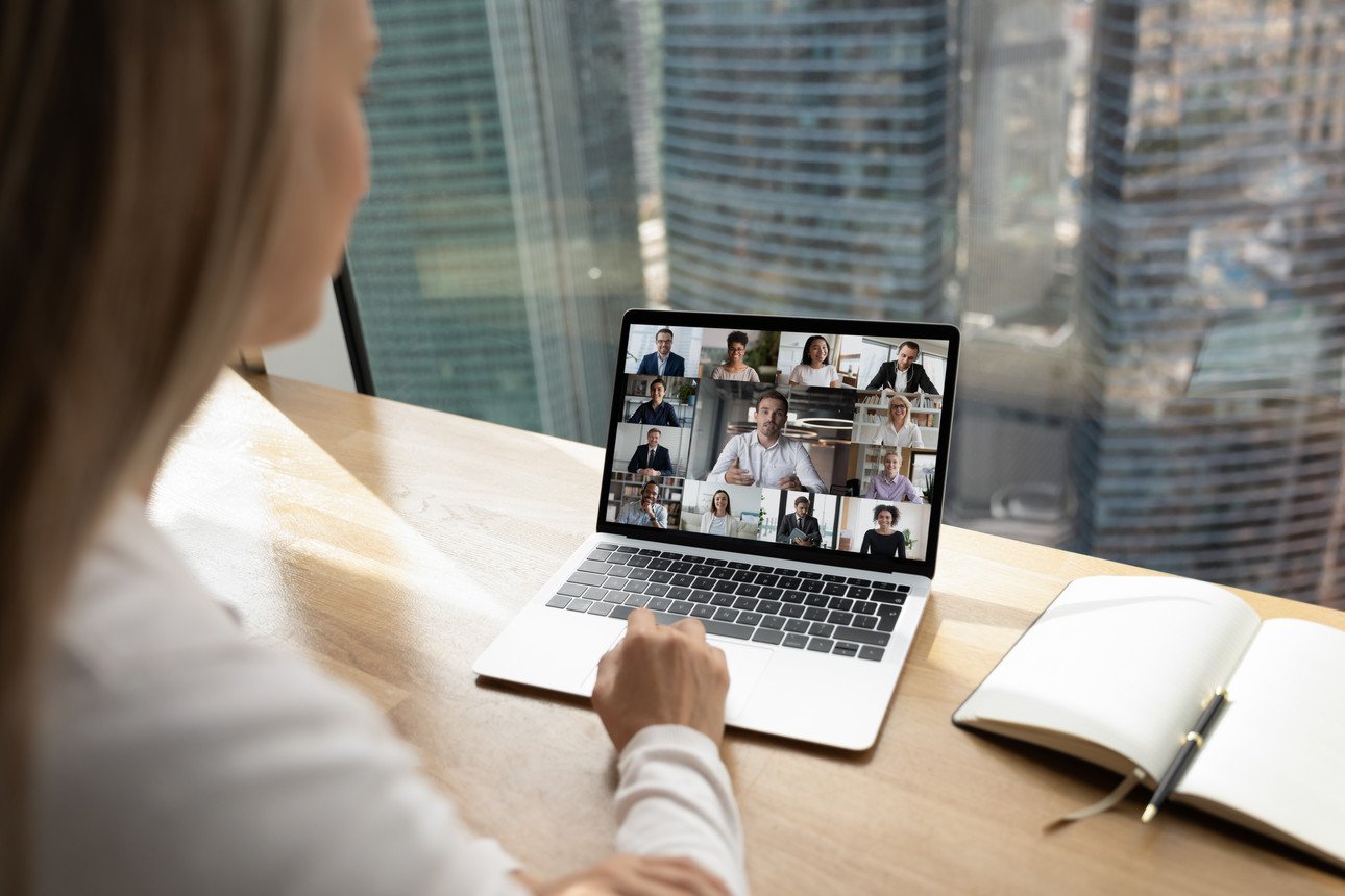 A blonde woman in a white blouse sits at a table and looks at a laptop on which participants of a video conference can be seen. 
