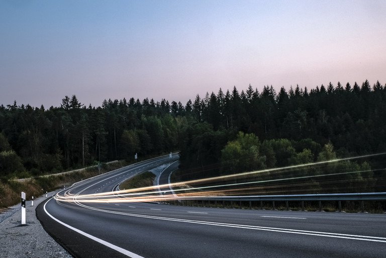 Symbolbild f&uuml;r Mobilit&auml;t im l&auml;ndlichen Raum. Autolichter auf einer l&auml;ndlichen Stra&szlig;e, umgeben von Wald.