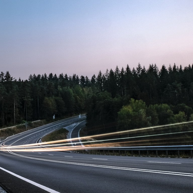 Symbolbild f&uuml;r Mobilit&auml;t im l&auml;ndlichen Raum. Autolichter auf einer l&auml;ndlichen Stra&szlig;e, umgeben von Wald.