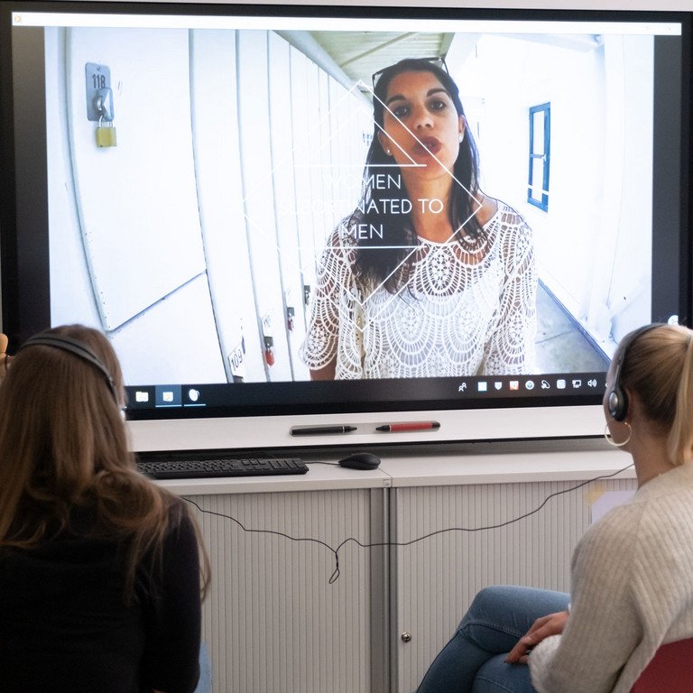 Aufnahme von zwei Frauen, die sich auf einer Leinwand die Video-Installation zum Thema sexualisierte Gewalt gegen Frauen in Popul&auml;rkultur ansehen.