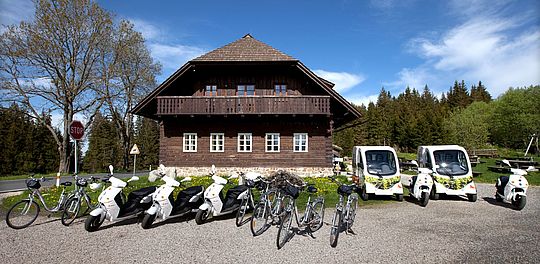 Elektrofahrzeuge vor einem alten Holzhaus im Böhmerwald. Foto: e-Sumava