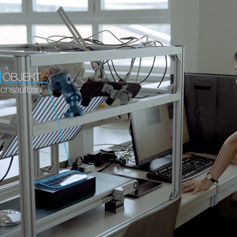 Eine Forscherin st&uuml;tzt sich auf einen Tisch mit einem wissenschaftlichen Aufbau, bei dem ein spiegelndes Objekt unter einer Kamera vermessen werden soll. Sie schaut in den Computer.