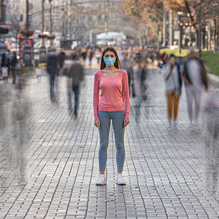 Frau mit Mundschutz steht auf einer belebten Stra&szlig;e, blickt in die Kamera, andere Menschen verschwinden in der Bewegungsunsch&auml;rfe. Symbolbild: Adobe Stock