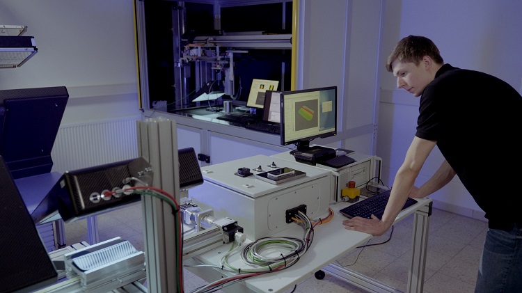 Ein junger Forscher lehnt an einem Tisch mit einem wissenschaftlichen Aufbau, auf dem ein spiegelndes Objekt auf einem Flie&szlig;band vermessen werden soll. 