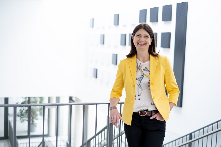 Frau Professorin Dr. Carolin H&auml;ussler, eine gro&szlig;e Frau mit langem, braunen Haar, lehnt l&auml;ssig an einem Gel&auml;nder. Sie tr&auml;gt einen strahlend gelben Blazer und l&auml;chelt freundlich in die Kamera. 
